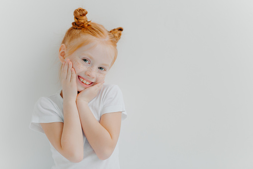 Shy pretty girl has two hair buns, touches cheeks gently, smiles happily, has freckles, hears something positive from parents, wears white t shirt, stands indoor, blank space left for your information