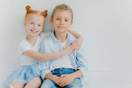 Friendly small girl and boy hug and sit on floor against white background, have positive expressions, feel satisfied after playing together, copy space. Ginger female kid embraces brother with love