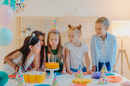 Small kids celebrate birthday party, blow candles on cake, gather at festive table, have good mood, enoy spending time together, make wish, wear party hats, pose indoor with inflated balloons