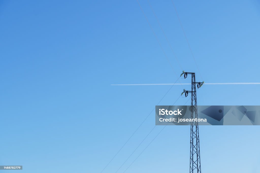 high tension tower in the background blue sky and an airplane passing by leaving its wake a plane passes leaving its wake behind a high tension tower, in the background a clear blue sky Aerial View Stock Photo