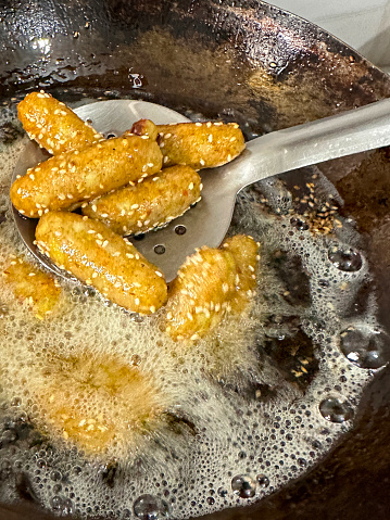 Stock photo showing close-up, elevated view of a wok full of bubbling, boiling oil used to deep fry a pile of golden brown, homemade, sesame seed coated, potato croquettes.