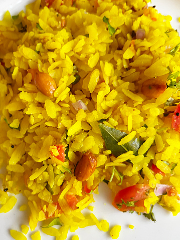 Stock photo showing close-up, elevated view of poha rice dish of spiced flattened rice with onion, green chillies, curry and coriander leaves, peanuts, mustard and cumin seeds, turmeric and lemon juice on a white plate.