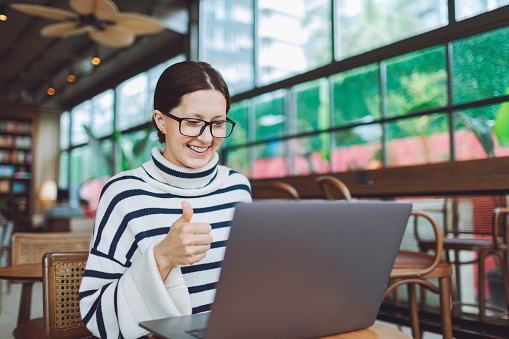 Young woman in a cafe smiles while working on laptop and giving a thumbs up during video call with colleagues