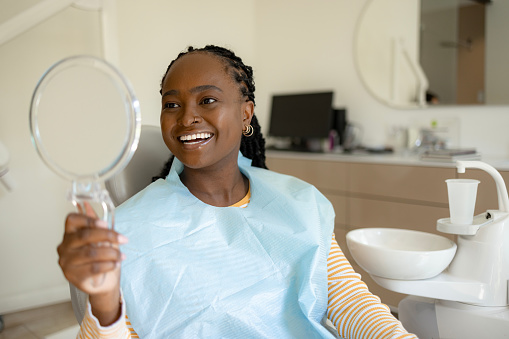 Happy African American woman using hand mirror to look at her clean teeth at the dentists'.