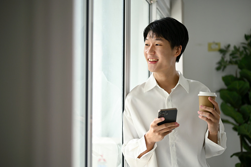 Happy and smiling young Asian man with his smartphone and coffee cup, looking out and leaning on the window, and daydreaming about his career.