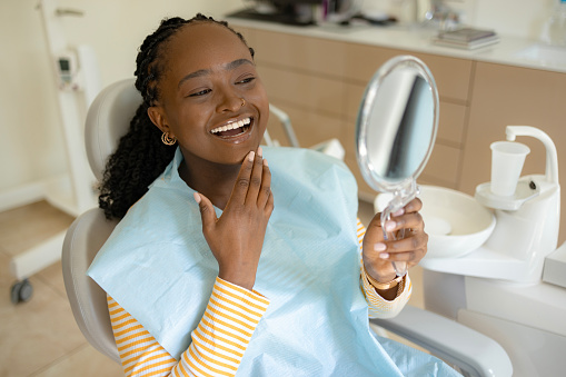 Happy African American female patient looking at her whitened teeth in hand mirror at the dentists'.