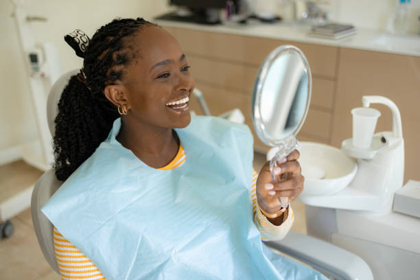 Black woman feeling happy while looking at her teeth in a mirror at the dentists'.