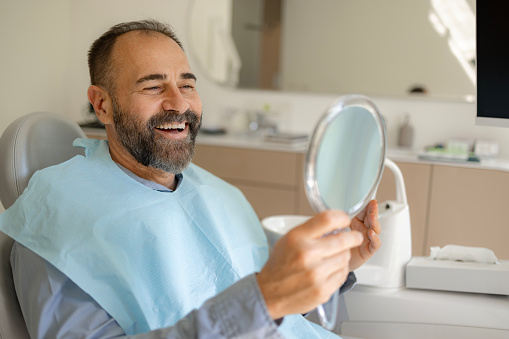 Mid adult man feeling happy while looking at his clean teeth in a mirror at the dentist's office.