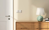 Close up of mid century style wooden sideboard with pastel green geometric shape of lamp shade, books in sunlight from window to cream wall with light switch and white door at entrance hall