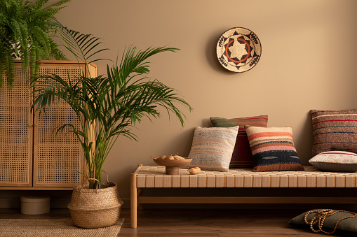 Warm and cozy ethno living room interior with couch, patterned pillows, plants i flowerpots, fern, rattan sideboard, basket on wall, wooden bowl and personal accessories. Home decor. Template.