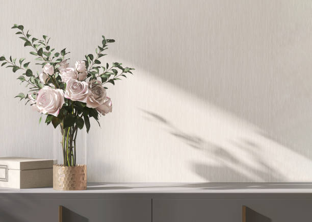 モダンでミニマルなグレーのキャビネットの空きスペース、ピンクのバラの花束とガラスの花瓶、日光の当たる金のアクセサリーボックス、空白のベージュの壁紙壁に影