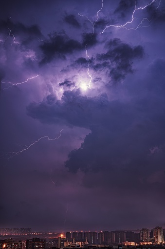 A dramatic view of a storm illuminating the night sky with flashes of lightning, against a backdrop of illuminated buildings in the distance