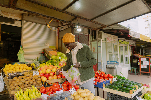 Photo of a young African American man buying fruits and vegetables from a street market stand, choosing healthy snack for the day. He is doing weekly shopping and running errands on his cargo bike.