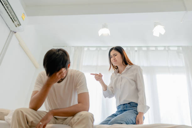 violenza domestica, moglie asiatica che minaccia il marito, leader della famiglia spaventato che abbraccia il suo mentre è seduto insieme sul letto. - bullying family violence domination foto e immagini stock