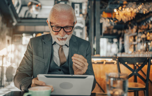 istock Mature business man, elegantly dressed, sitting in a coffee shop, focused and engaged while working online on a digital tablet, talking to clients or reading emails 1488809810