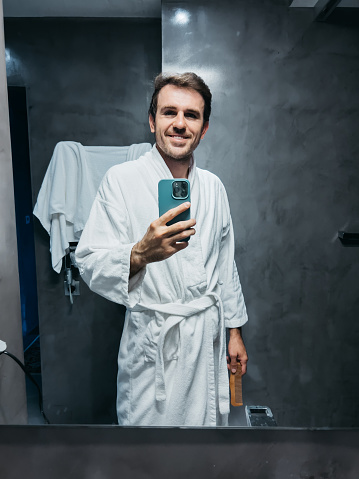 Smiling young man in white coat taking selfie with smartphone near bathroom mirror. High quality photo