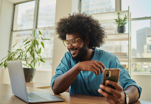 Happy, young creative business man with an afro, sitting at a desk in a relaxed office space, wearing a blue shirt whist holding a mobile cell phone and looking a laptop computer.