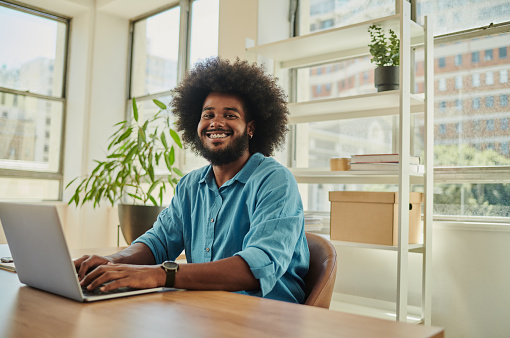 Happy, young black man wearing a blue shirt with an afro, smiling with dental braces whilst sitting at his desk using a laptop computer. Stock photo with copy space