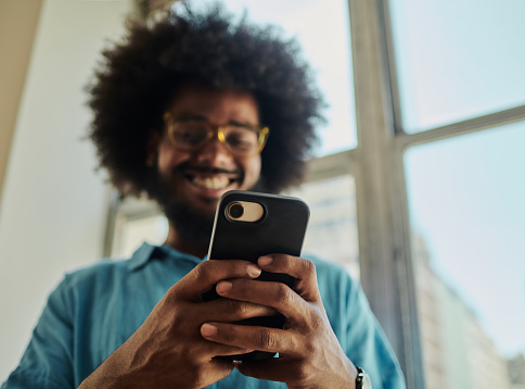 Defocused shot of a happy young black man with an afro, beard and dental braces standing in front of a  window smiling wearing a blue shirt, and eye glasses whilst holding his mobile phone to text stock photo