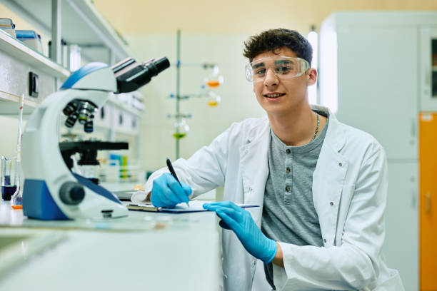 Happy biochemistry student taking notes during lab research and looking at camera. Young scientist writing notes while working on new research in laboratory and looking at camera. biochemist stock pictures, royalty-free photos & images
