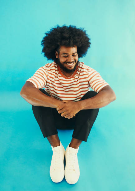 istock Young black man with a afro sitting on the ground or floor smiling, looking down feeling content whilst being dressed casually shot against a blue background stock photo 1488815808