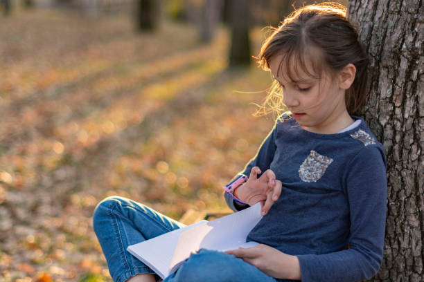 istock Child girl reading book while leaning on a tree in public park 1488815815