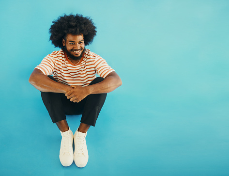 Young black bearded afro man sitting casually on the floor with his arms on his knees , smiling looking off camera shot against a blue background with copy space. stock photo