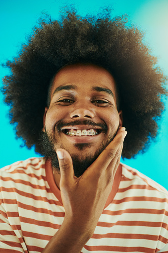 Young fresh faced black man with an afro smiling into camera with dental braces. Young man with his hand on his chin whilst wearing a striped t-shirt shot against a blue background stock photo