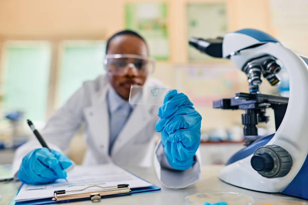 istock Close up of biochemist writing research data while examining sample on microscope slide in laboratory. 1488815857