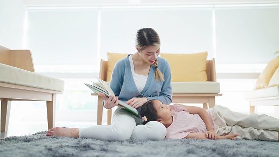 Asian mother reading a book to little daughter while listen on mother's lap on floor in living room. Family and love concept