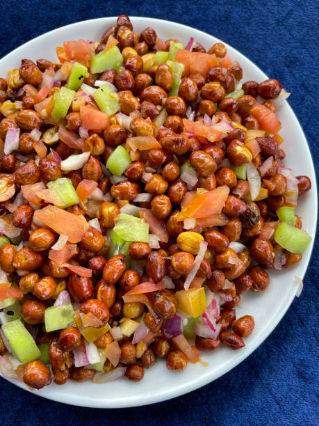 Stock photo showing elevated view of homemade peanut masala chaat recipe made with peanuts, chaat masala, chilli powder, salt, lemon juice, coriander, sliced serrano pepper (Capsicum annuum), diced tomato and red onion, served in a white bowl on blue velvet background. imagen de un tazón blanco que contiene masala chaat de maní casero, bocadillo salado al estilo callejero, cacahuetes, chaat masala, chile en polvo, sal, jugo de limón cilantro, chile serrano en rodajas (capsicum annuum), tomate cortado en cubitos y cebo