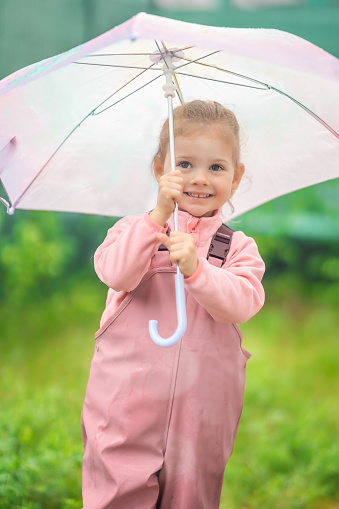 Happy laughing child girl 2-3 year old wearing waterproof clothes and holding pink umbrella have a fun on home backyard in rainy day. High quality photo
