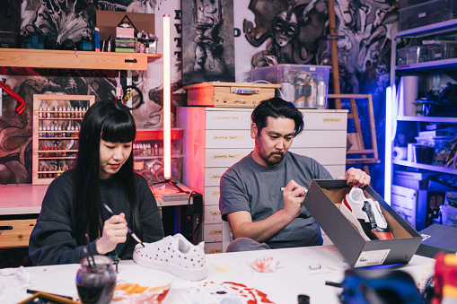 Two Japanese sneaker artists huddle over a workbench, discussing the finer points of their design as they carefully apply intricate details to a pair of custom sneakers.
