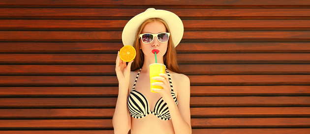 Summer portrait of happy young woman drinking fresh juice with slice of orange fruits wearing straw hat, bra