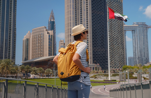 Enjoying travel in United Arabian Emirates. Young woman with yellow backpack walking on Dubai Downtown in sunny summer day. view from the back or rear view