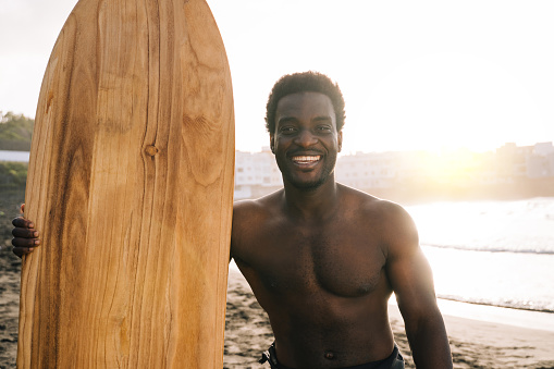Happy African surfer holding surf wood surfboard after riding waves on the beach - Sport training during summer vacation