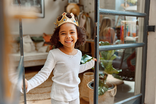 Crown, happy and portrait of child at a living room window smile, playing and having fun at home. Princess, game and face of girl relax, playful and carefree, curious and fantasy, imagine and joyful