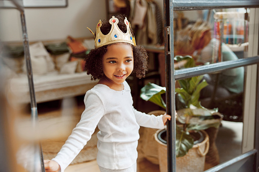 Crown, happy and child at a living room window smile, playing and having fun in her home. Princess, game and girl relax, playful and carefree, curious and fantasy, imagine and enjoying the weekend