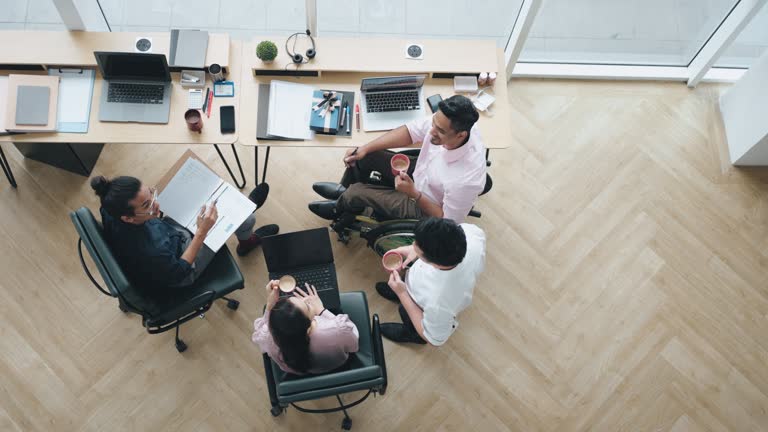 directly above Indian white collar male worker in wheelchair having cheerful discussion conversation with colleague in creative office workstation beside window