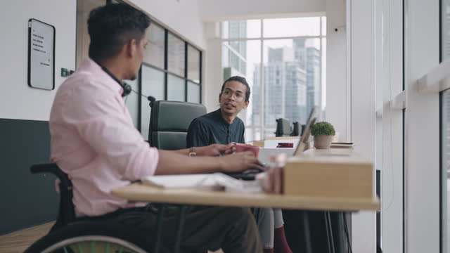 Asian Malay man talking to his Indian colleague in wheelchair brainstorming at workplace