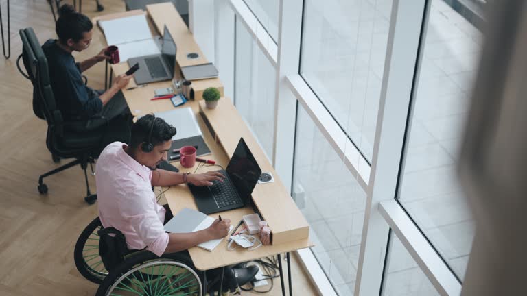 Asian Indian white collar male worker in wheelchair concentrating working in office beside his colleague