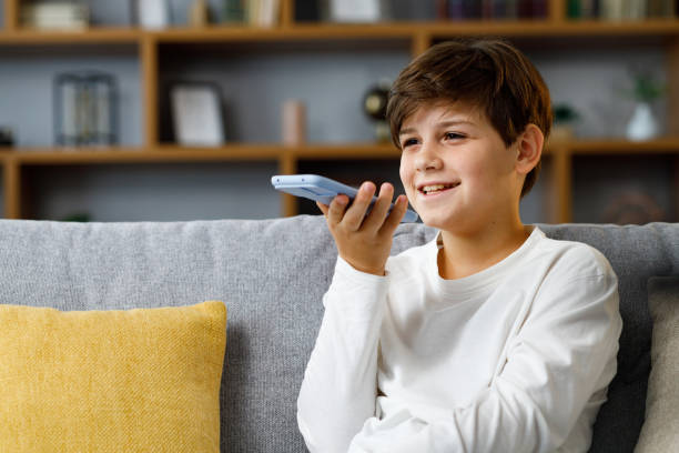 giovane ragazzo sveglio che registra il messaggio vocale sullo smartphone. adolescente sorridente che utilizza l'app di assistente virtuale seduto all'allenatore a casa. riconoscimento vocale, messaggi audio, utilizzo del traduttore sul telefono cellulare - one little boy audio foto e immagini stock