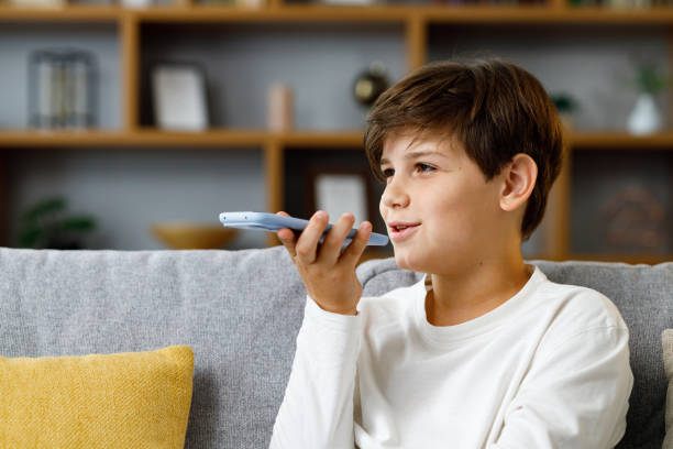 giovane ragazzo sveglio che registra il messaggio vocale sullo smartphone. adolescente sorridente che utilizza l'app di assistente virtuale seduto all'allenatore a casa. riconoscimento vocale, messaggi audio, utilizzo del traduttore sul telefono cellulare - one little boy audio foto e immagini stock