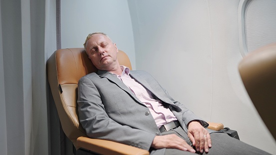 Exhausted middle aged caucasian businessman in suit sleeping on comfortable seat of private business jet. The passenger is sleeping while travel by aircraft