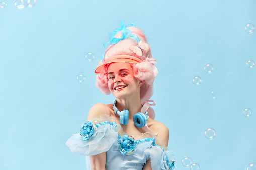 Close up portrait of adorable, young princess, queen with soap bubbles smiling at camera with happy face over blue background. Concept of comparison of eras, baroque style, beauty, fashion, emotion