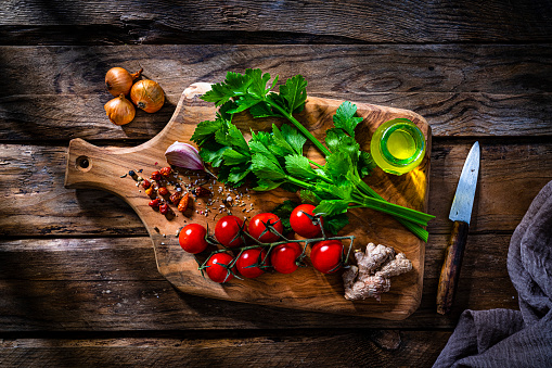 Cutting board with fresh vegetables and spices ingredients shot from above on rustic wooden table. The composition includes onion, ginger, peppercorns, tomato, garlic, celery and extra virgin olive oil bottle. High resolution 42Mp studio digital capture taken with SONY A7rII and Zeiss Batis 40mm F2.0 CF lens