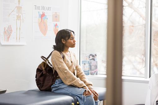 The young adult female patient gazes out the window while waiting for the doctor to come into the room.