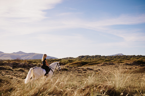 Beautiful young blond woman riding out with her white horse between the green sand dunes and under the blue sky. Color editing. Part of a series.