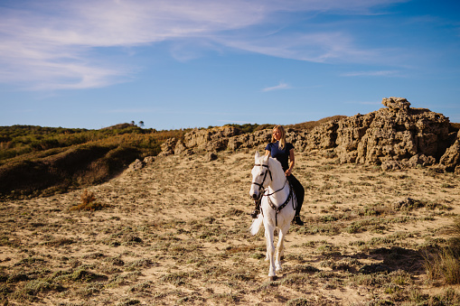 Beautiful young blond woman riding out with her white horse between the green sand dunes and under the blue sky and in front of sandstone rocks. Color editing. Part of a series.