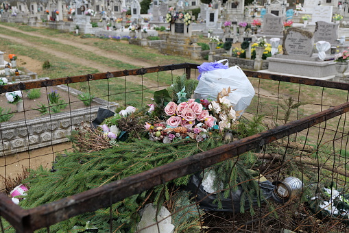 Rusty wire mesh container bin in a cemetery full of decayed and plastic flowers, pine branches and other garbage. The picture was made in Hungary, in the village cemetery of Penészlek.